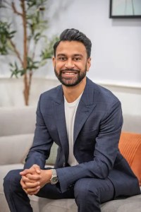 Professional portrait of a man in a dark blue suit seated in a modern office waiting area with contemporary decor and plants